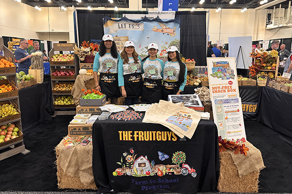 The FruitGuys Farm-to-School Team posing in their CSNA booth surrounded by fresh produce