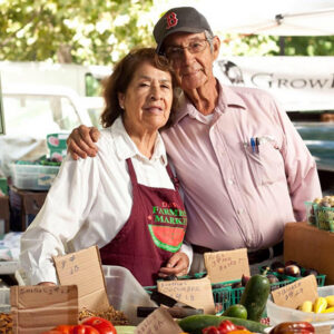 Smiling couple behind the table of a farmers market stand, surrounded by fresh fruits and vegetables