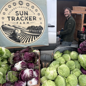 Smiling farmer near sun Tracker Farm sign, behind a table of fresh cabbages